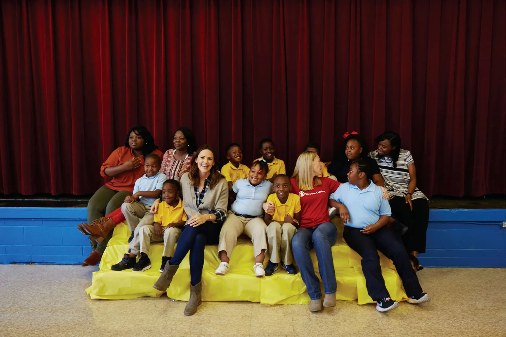 Group of diverse children and adults posing and smiling on a yellow platform in front of a red stage curtain.