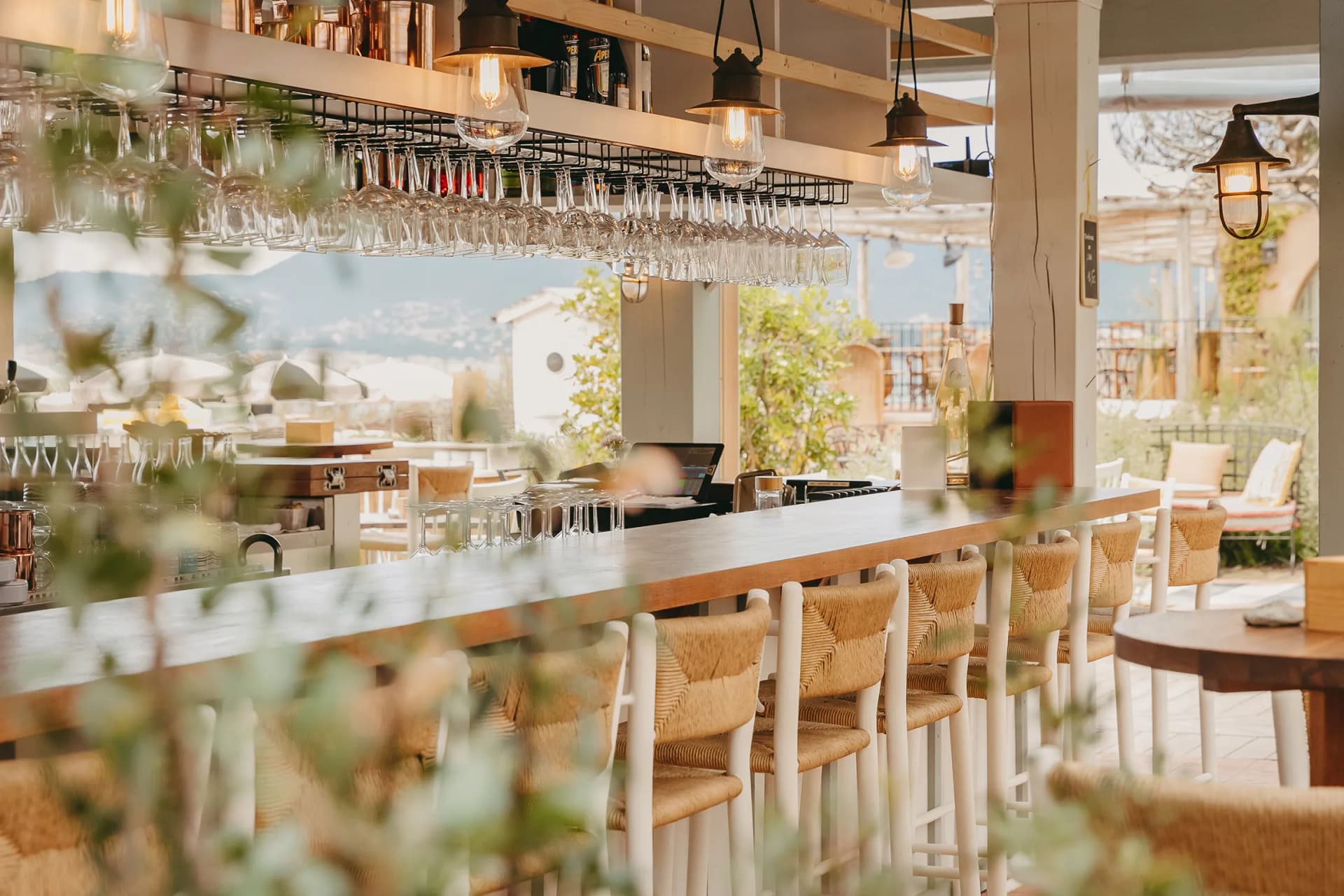 Outdoor bar with hanging wine glasses above a wooden counter, warm pendant lights, wicker stools, and distant mountains.