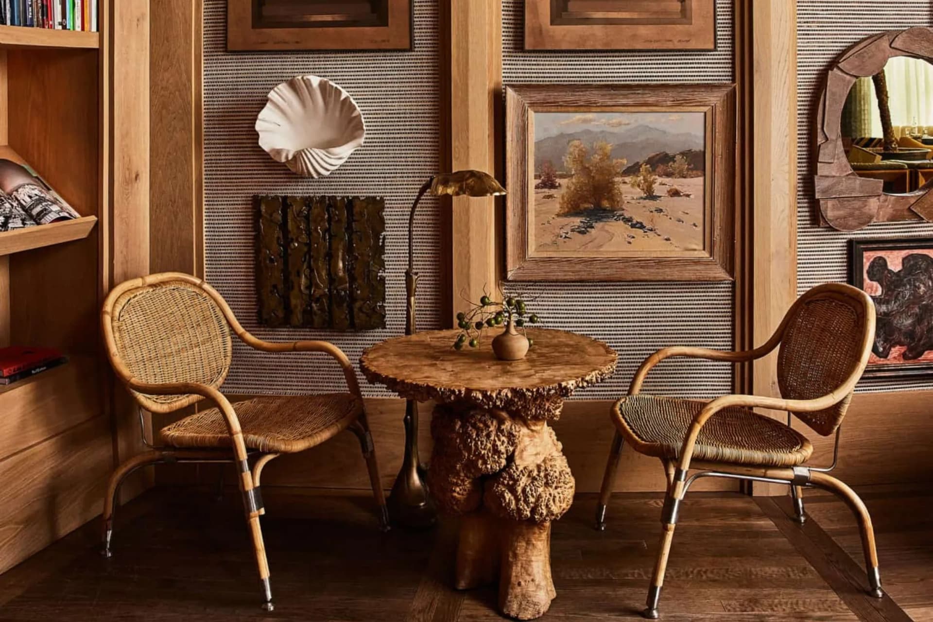 Rustic room with two wicker chairs around a carved wooden table; framed landscape paintings and a seashell wall sculpture.
