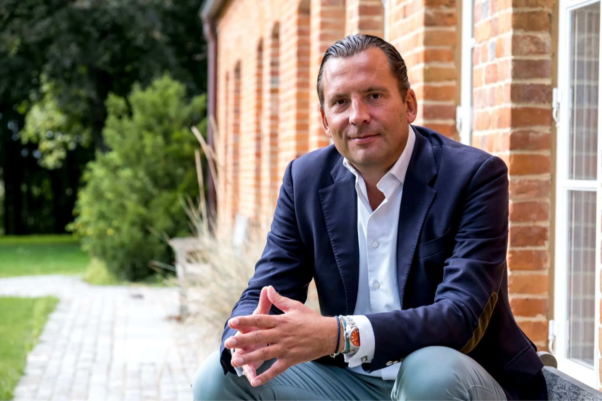 Man in a navy blazer and white shirt sits on brick steps outside, hands clasped, with greenery in the background.