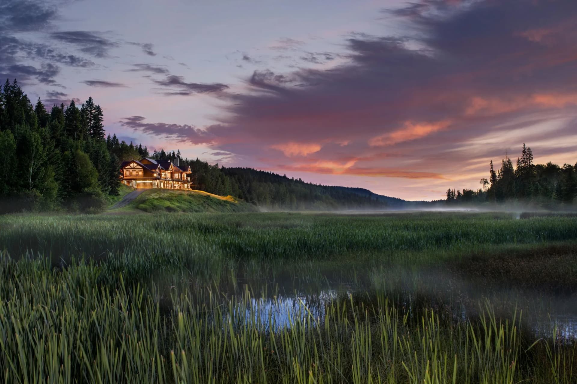 A warmly lit wooden hut on a hill at the edge of a pine forest, with mist-shrouded grass in the foreground and a pink sunset sky.