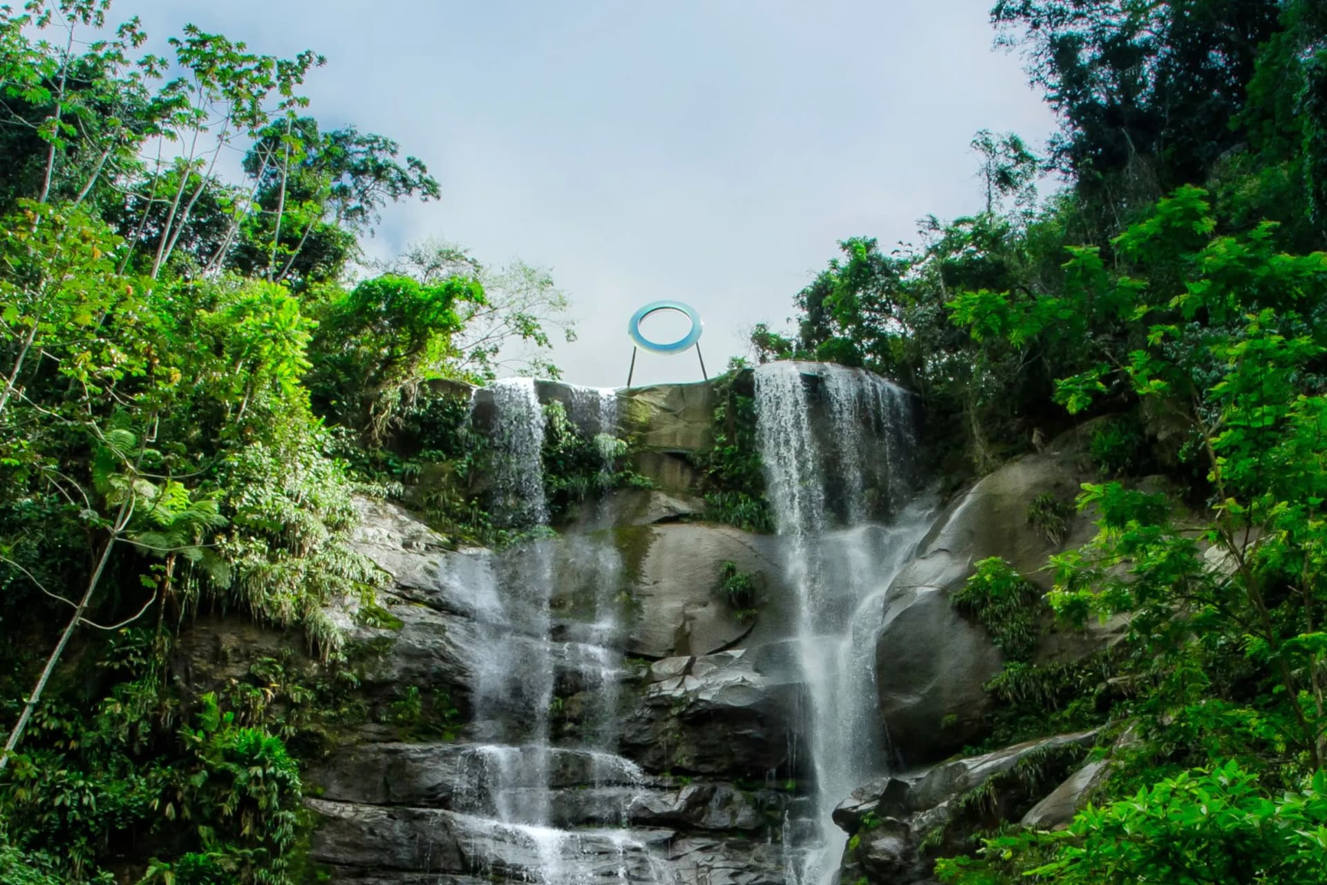 A waterfall cascades down rocky ledges amidst dense green jungle; a blue circular ring floats above the falls.