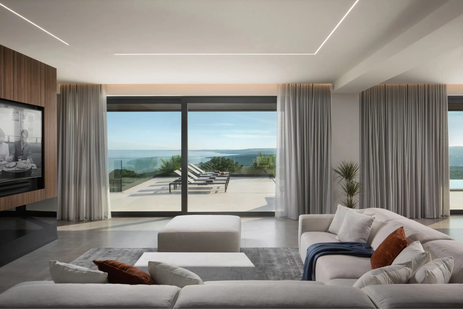 Modern living room with large glass doors opening to an ocean view, white sectional sofa, gray curtains, and a wall-mounted TV.