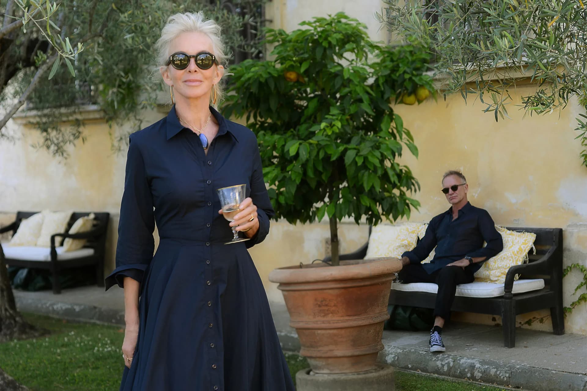 Elegant older woman in a navy dress and round sunglasses holds a wine glass outdoors; a man in dark clothes sits on a bench behind her amid olive trees.