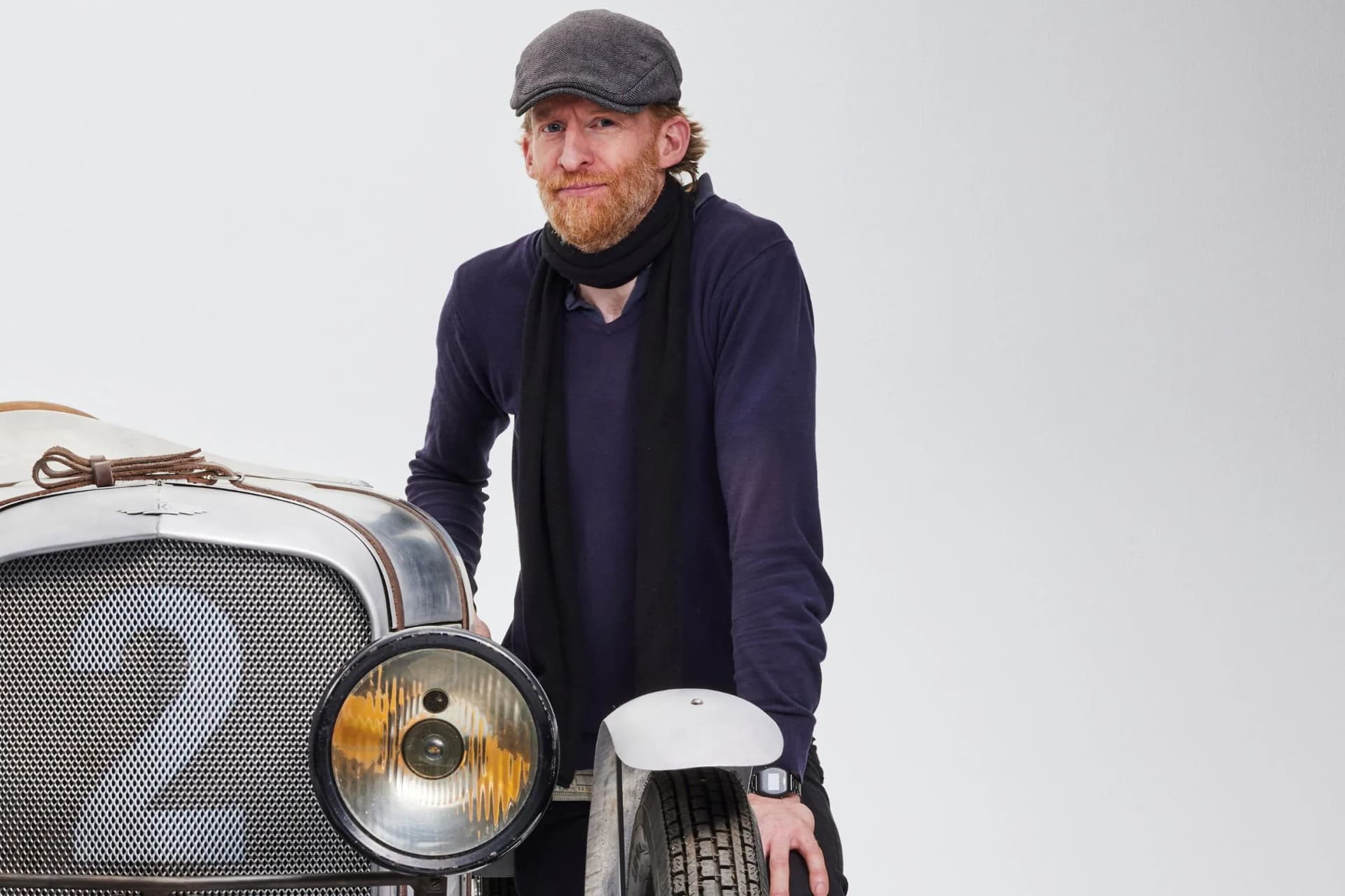 Bearded man wearing a gray cap and scarf leans on a vintage silver car with a round headlight, against a plain light background.
