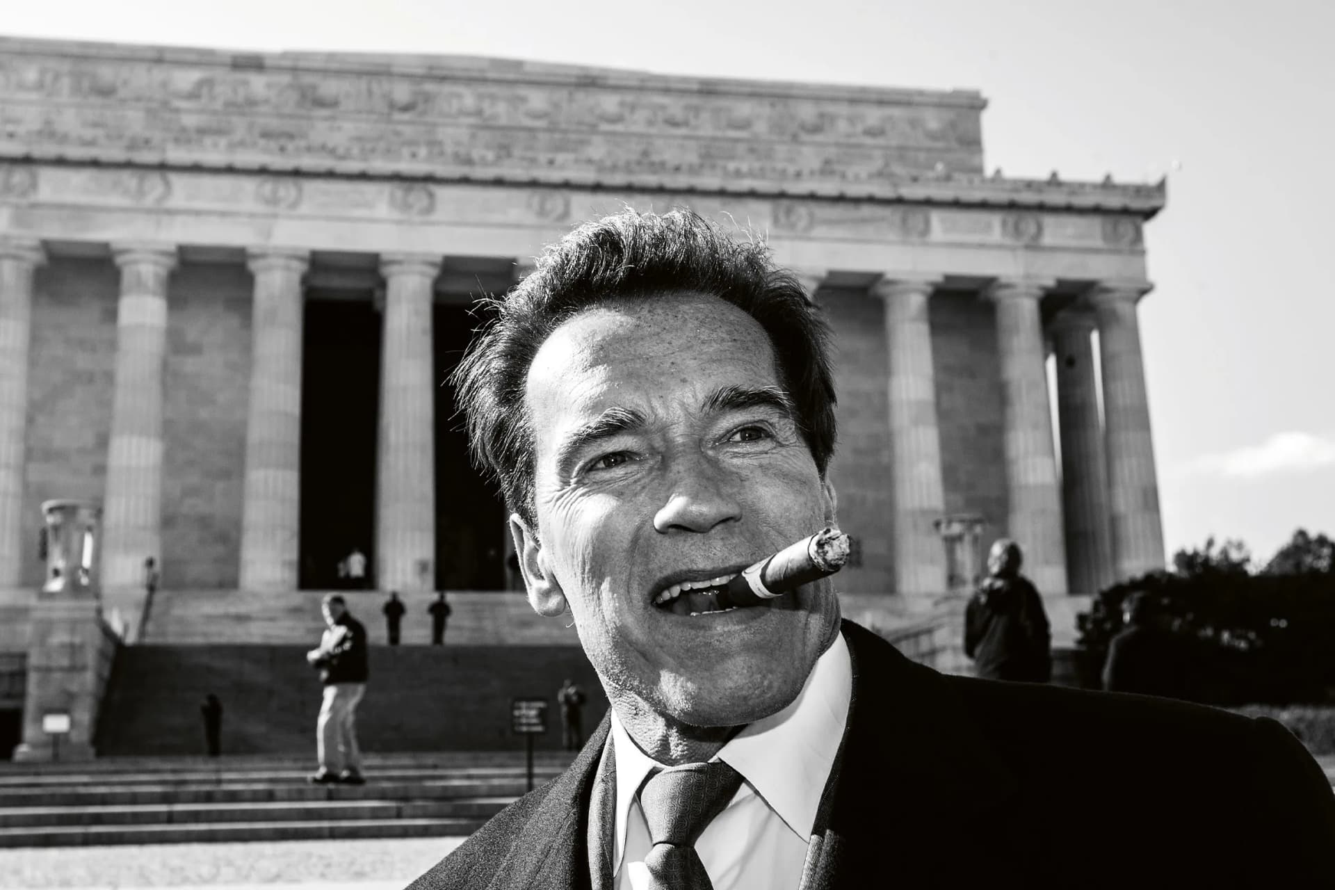 Black-and-white photo of a man in a suit smoking a cigar, standing in front of a large neoclassical building with tall columns.