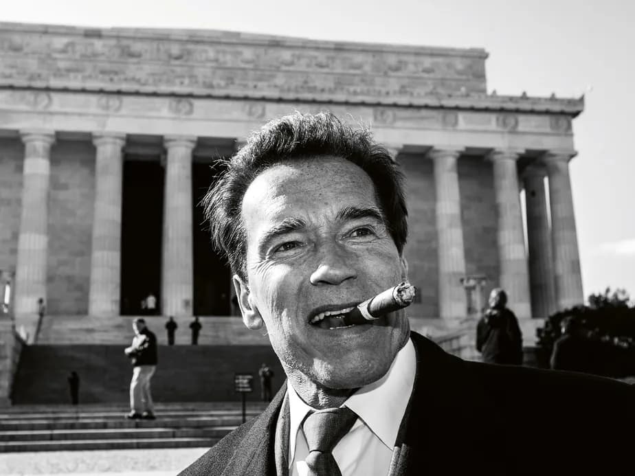 A black-and-white close-up of a smiling man in a suit, holding a cigar, in front of a neoclassical building with tall columns.