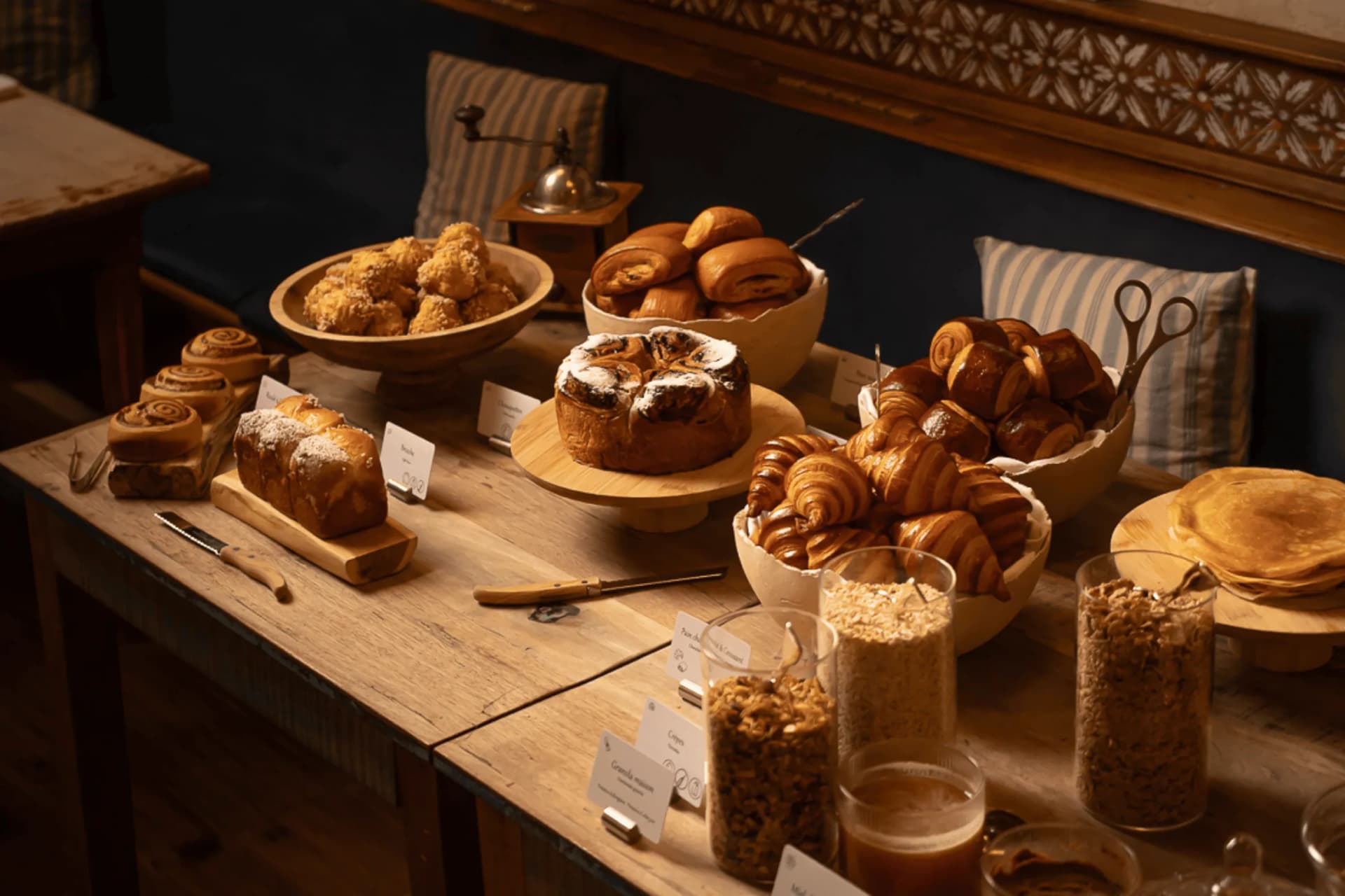 Rustikale Bäckerei-Auslage auf einem Holztisch: Croissants, Zimtschnecken, Brötchen und Gläser mit Müsli, unter warmer Beleuchtung und vor einer gemusterten Wand im Hintergrund.