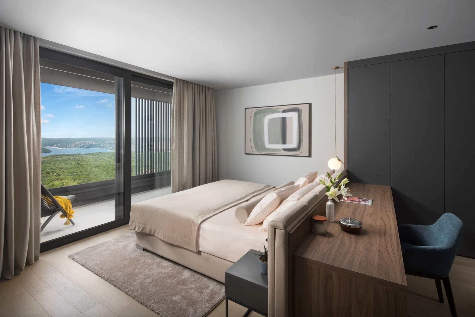 Modern bedroom with a large window and balcony view of hills and a lake; beige bed, abstract art, and a dark wood desk beside gray wardrobes.
