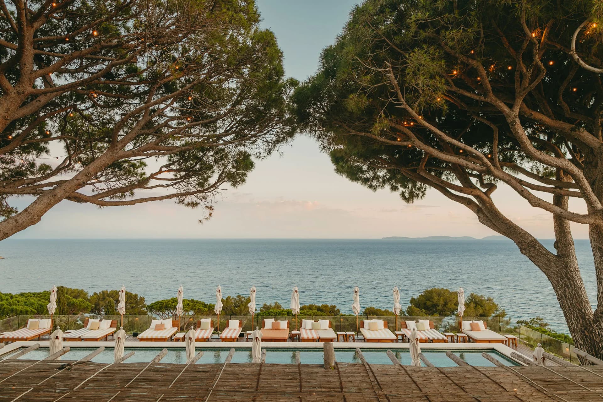 Rows of striped sun loungers and umbrellas along a pool at a seaside resort, framed by large trees with string lights, overlooking the calm sea.