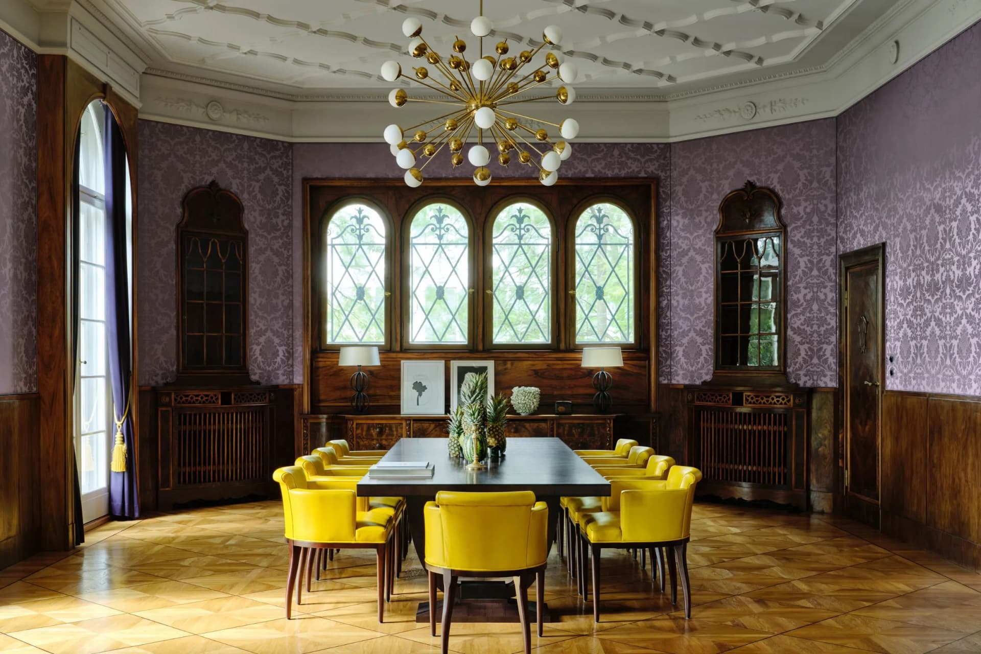 An elegant dining room with violet-patterned damask walls, arched windows, dark wood panelling and a golden star-shaped chandelier hanging above a long table with yellow chairs.