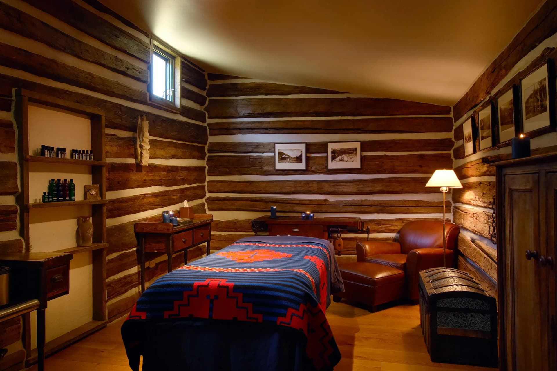 Cozy log cabin bedroom with rough-hewn wooden walls, a bed with a blue and red geometric quilt, leather chair, lamp, and framed art.