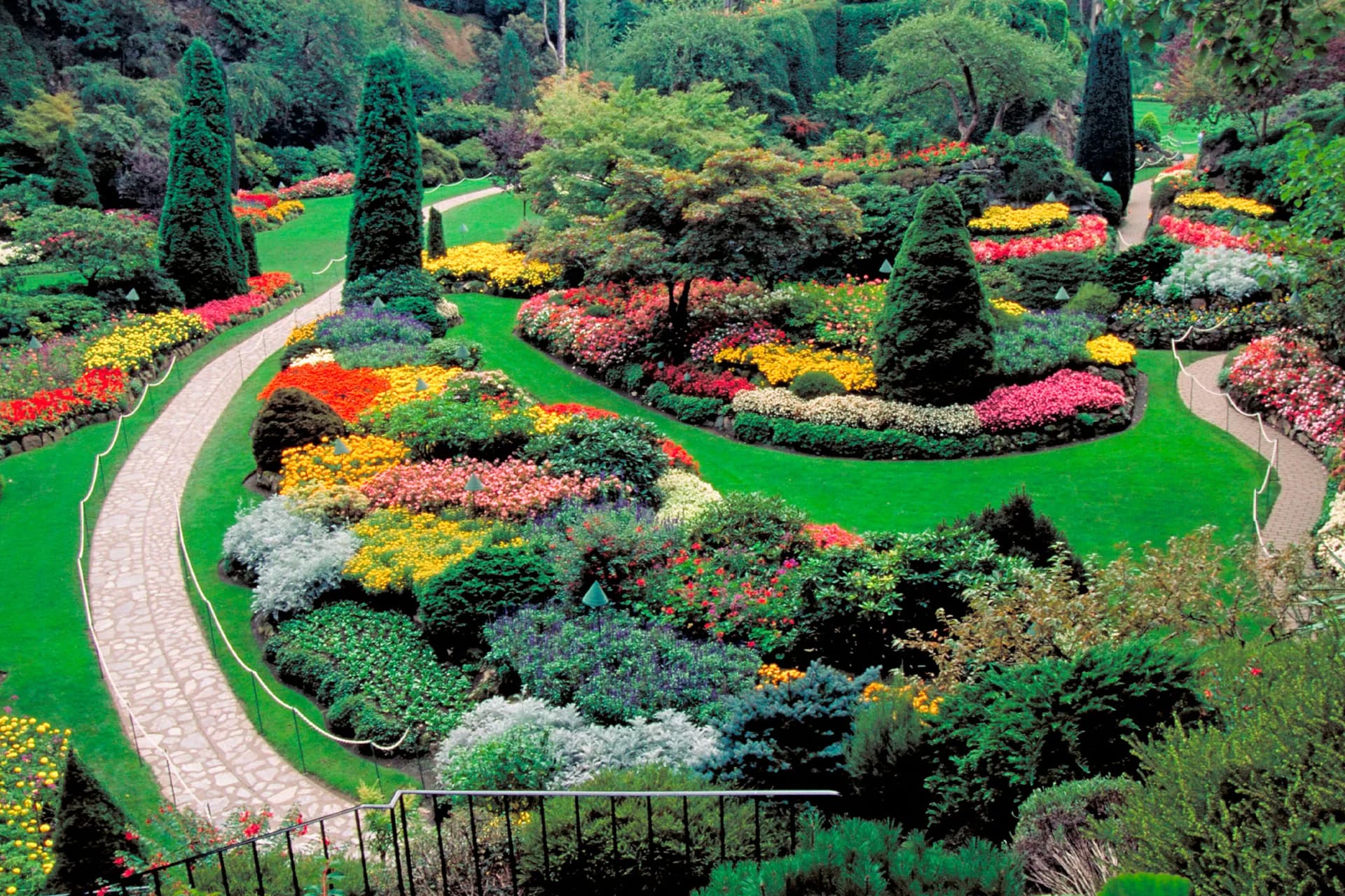 A vivid formal garden with winding stone paths, bright flower beds, and sculpted evergreen topiary amid lush green lawns.