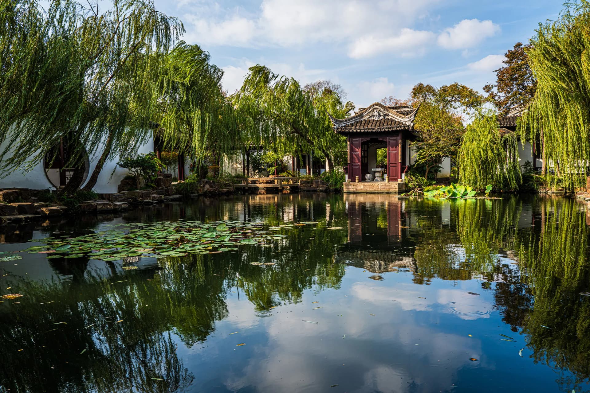 Tranquil Chinese garden pond framed by weeping willows, with an ornate curved-roof pavilion and lily pads, reflected in the water.