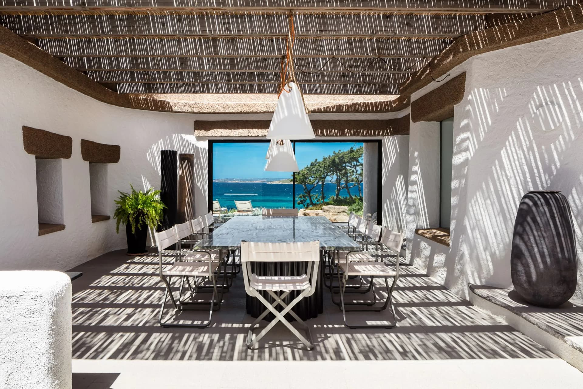 Open-air dining area with a long glass table and white chairs under a slatted wooden roof, opening to a sea-view with lounge chairs outside.