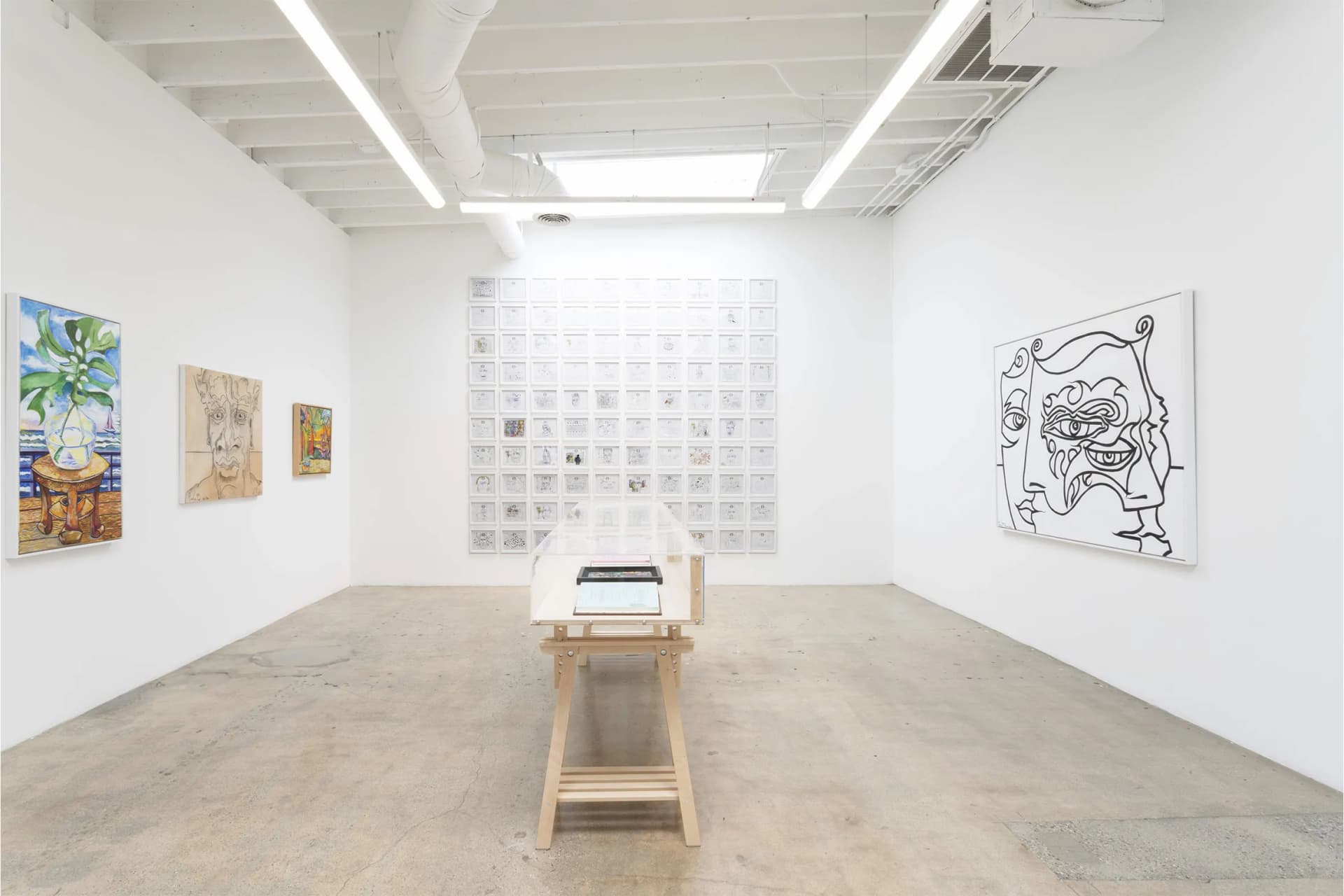 Bright white gallery with a wooden table; back wall of small framed pieces; left wall paintings; right wall black-and-white abstract drawing.