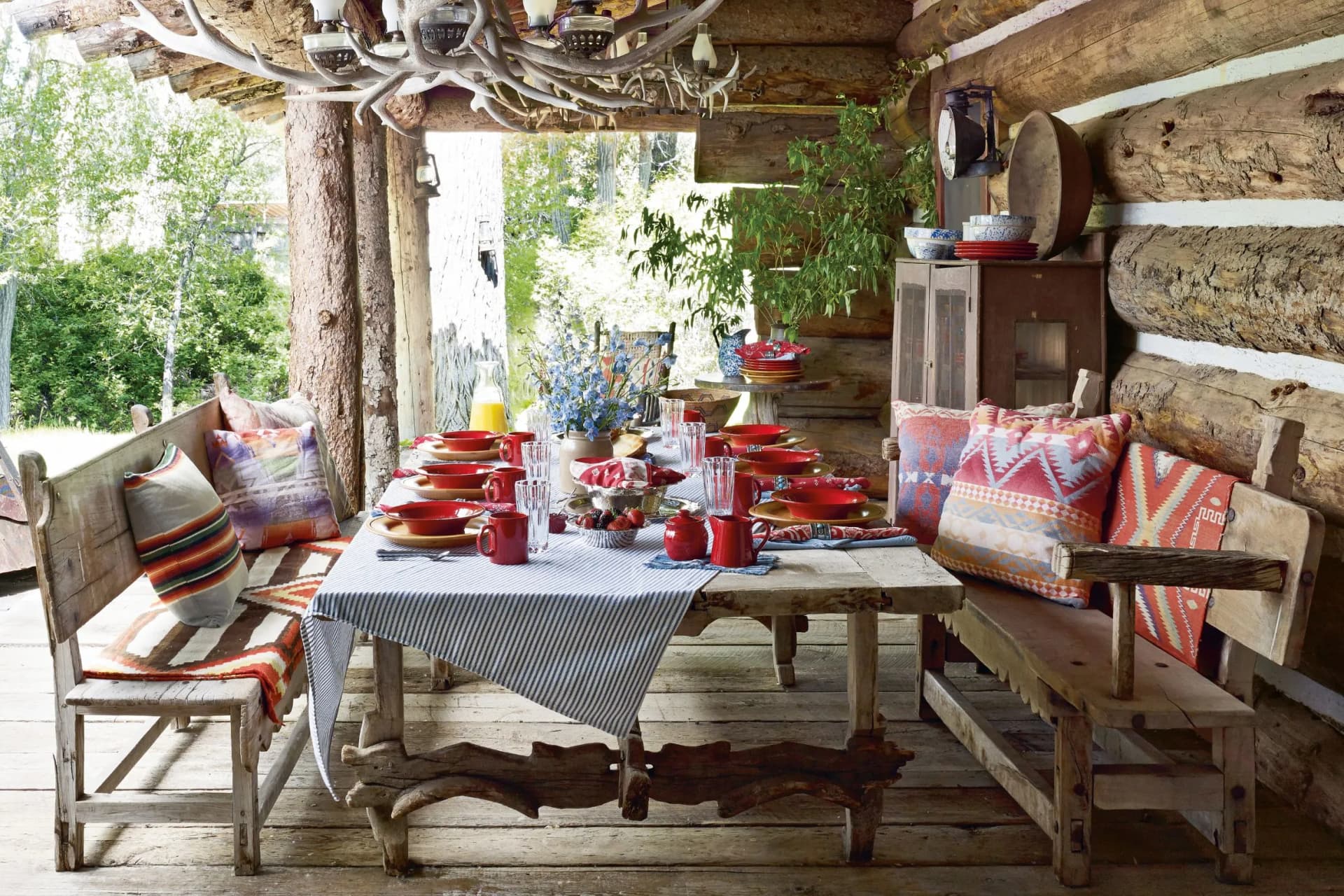 Rustic log cabin dining area with a long wooden table set for a meal, red mugs and plates, benches with colorful cushions, and an antler chandelier.