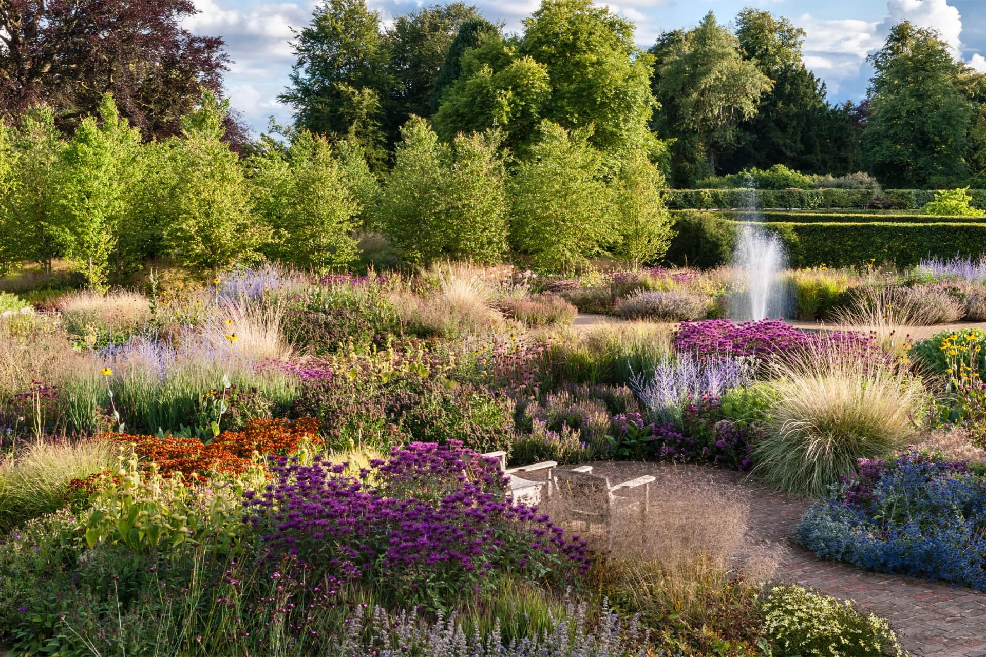 Sunlit garden with a central fountain, vibrant flower beds, ornamental grasses, and dense green trees in the background.
