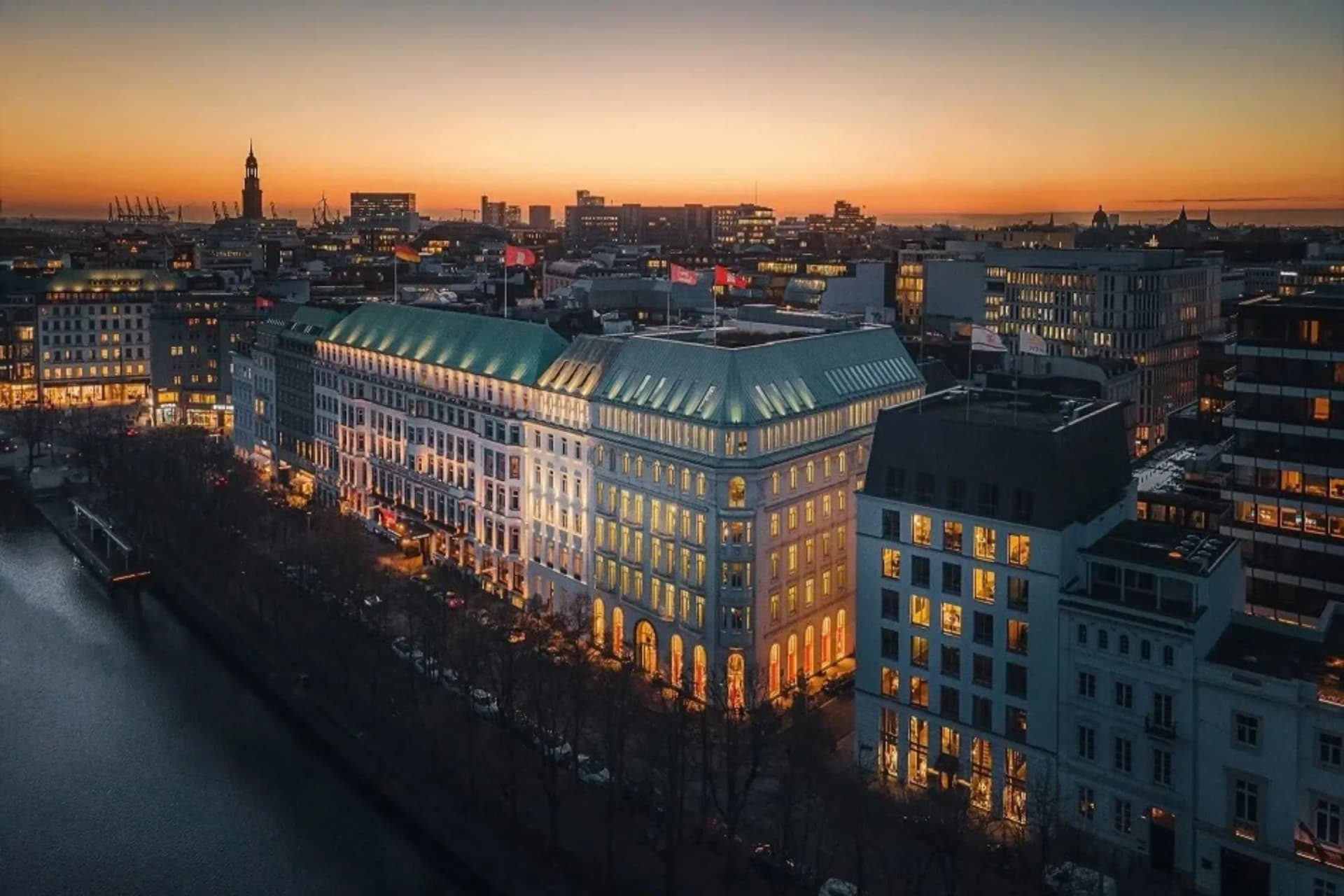 Tramonto sulla sagoma della città; in primo piano un edificio bianco con il tetto turchese e le finestre illuminate, sullo sfondo gru e grattacieli moderni.