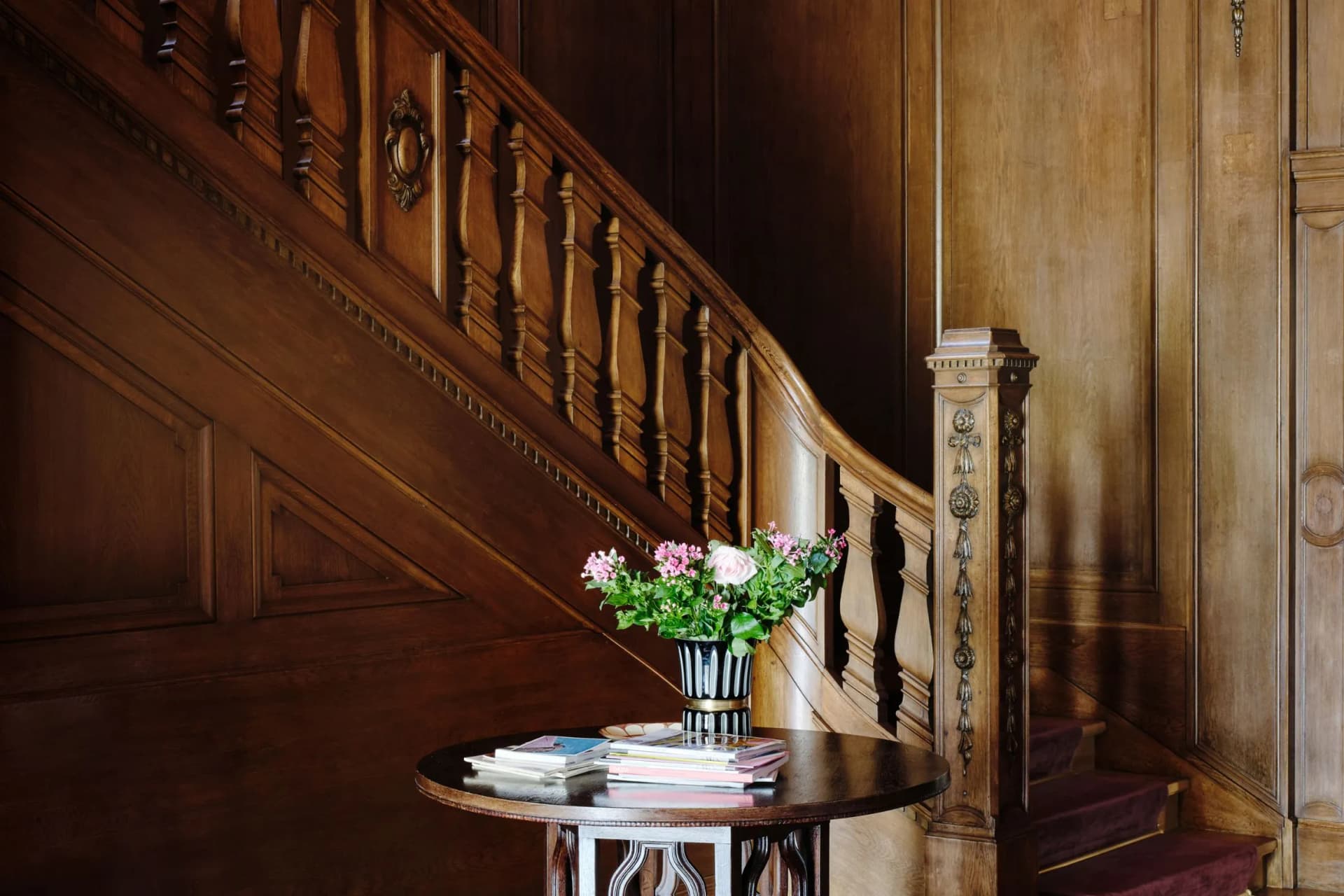 A dark wooden staircase with ornate banisters against panelled walls; on a round table stands a vase of pink and white flowers, along with some magazines.