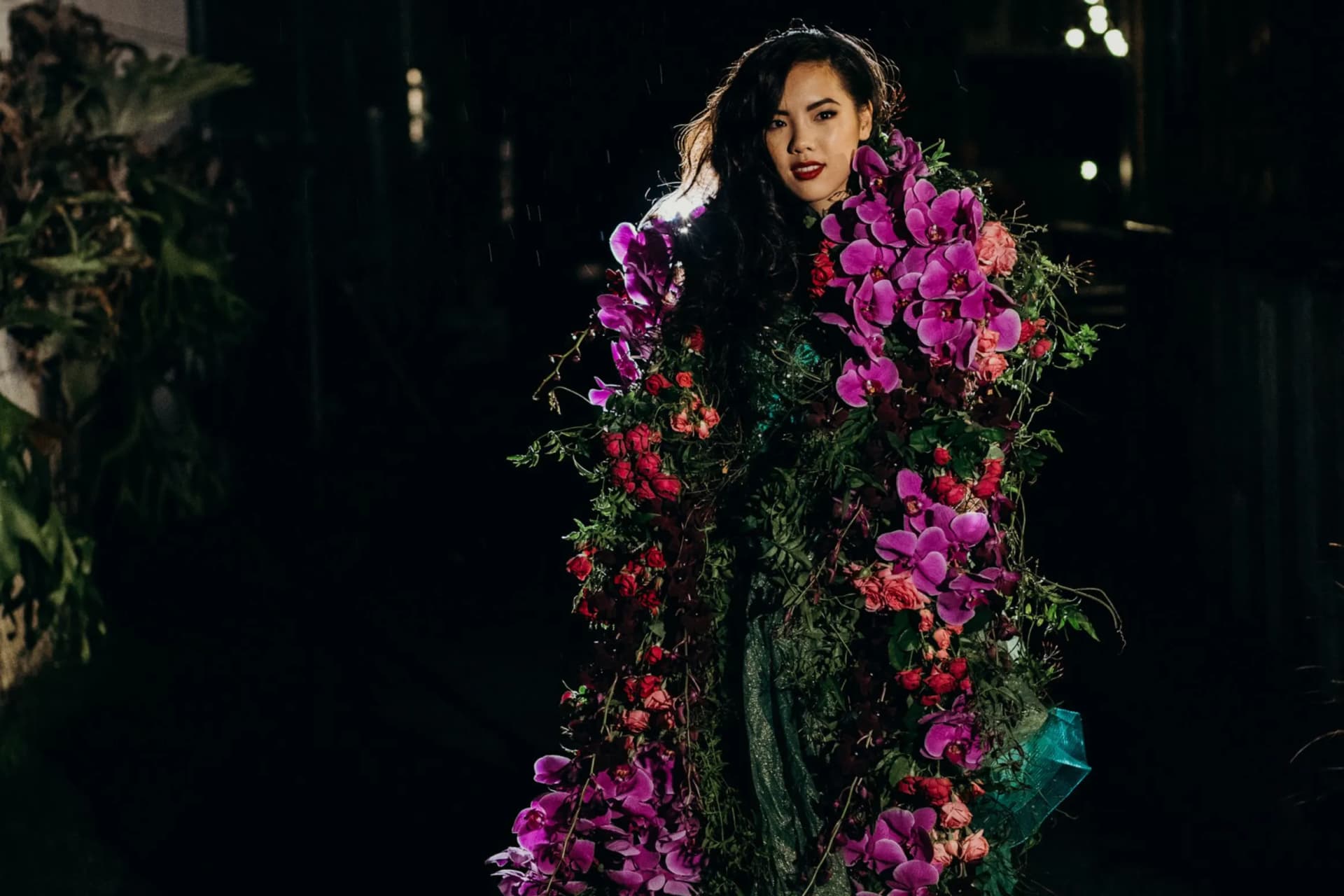 A woman stands in the dark, wrapped in a cascading bouquet of pink and purple orchids and red flowers.