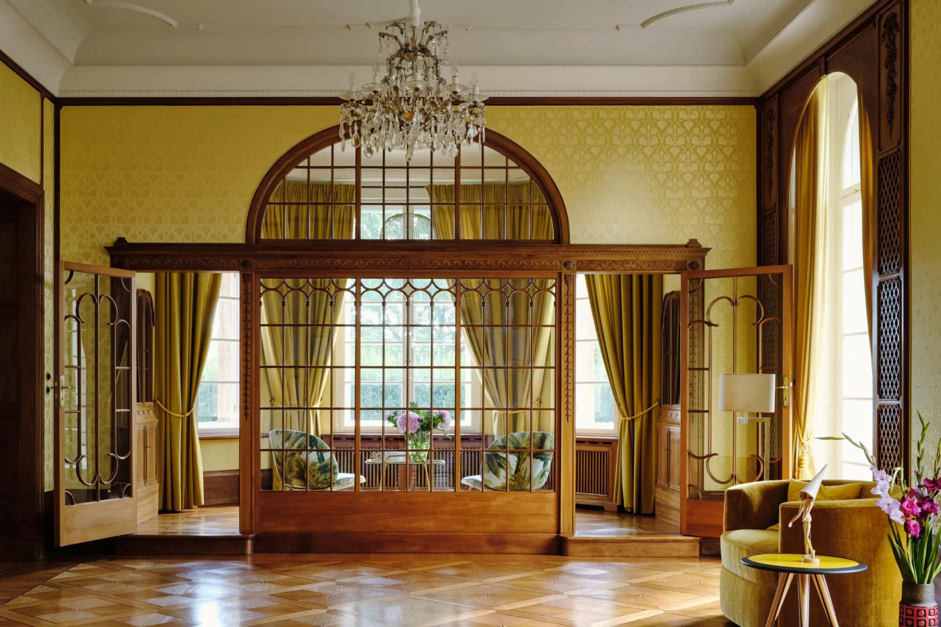 An elegant vintage room featuring ornate wood panelling, yellow damask walls, a large arched window, a crystal chandelier and gold curtains.
