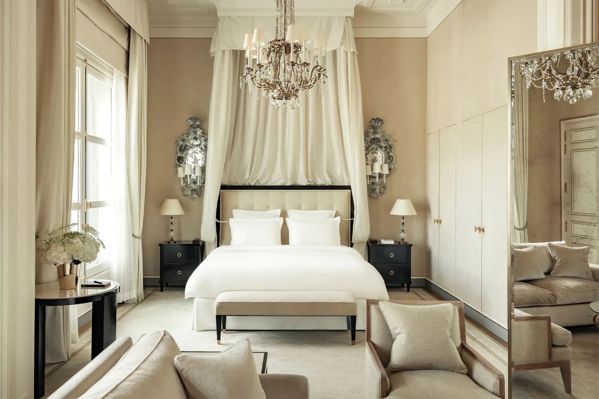 Luxurious bedroom with a white canopy bed, crystal chandelier, beige walls, flowing drapes, black nightstands, and a cozy seating area.