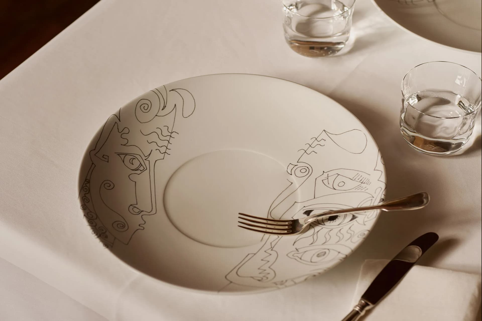 White plate with abstract line drawings of faces rests on a white tablecloth; a fork lies across its center, with a knife and glasses nearby.