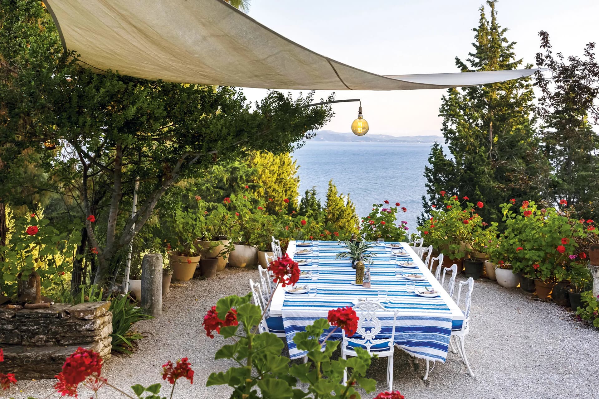 Seaside garden dining setup: long blue-and-white striped table, white chairs, shade sail, hanging bulb, potted geraniums, and distant blue sea.