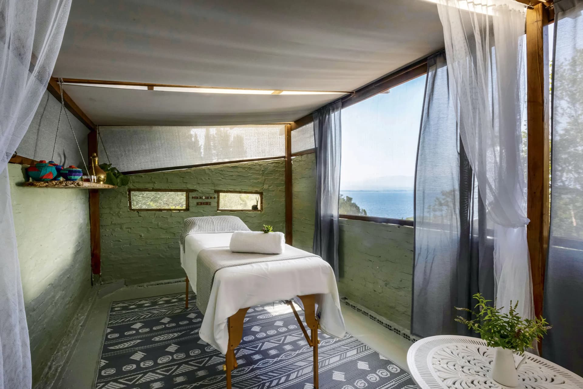 Sunlit spa alcove with a massage table under a fabric canopy, sheer curtains, brick walls, and an ocean view.