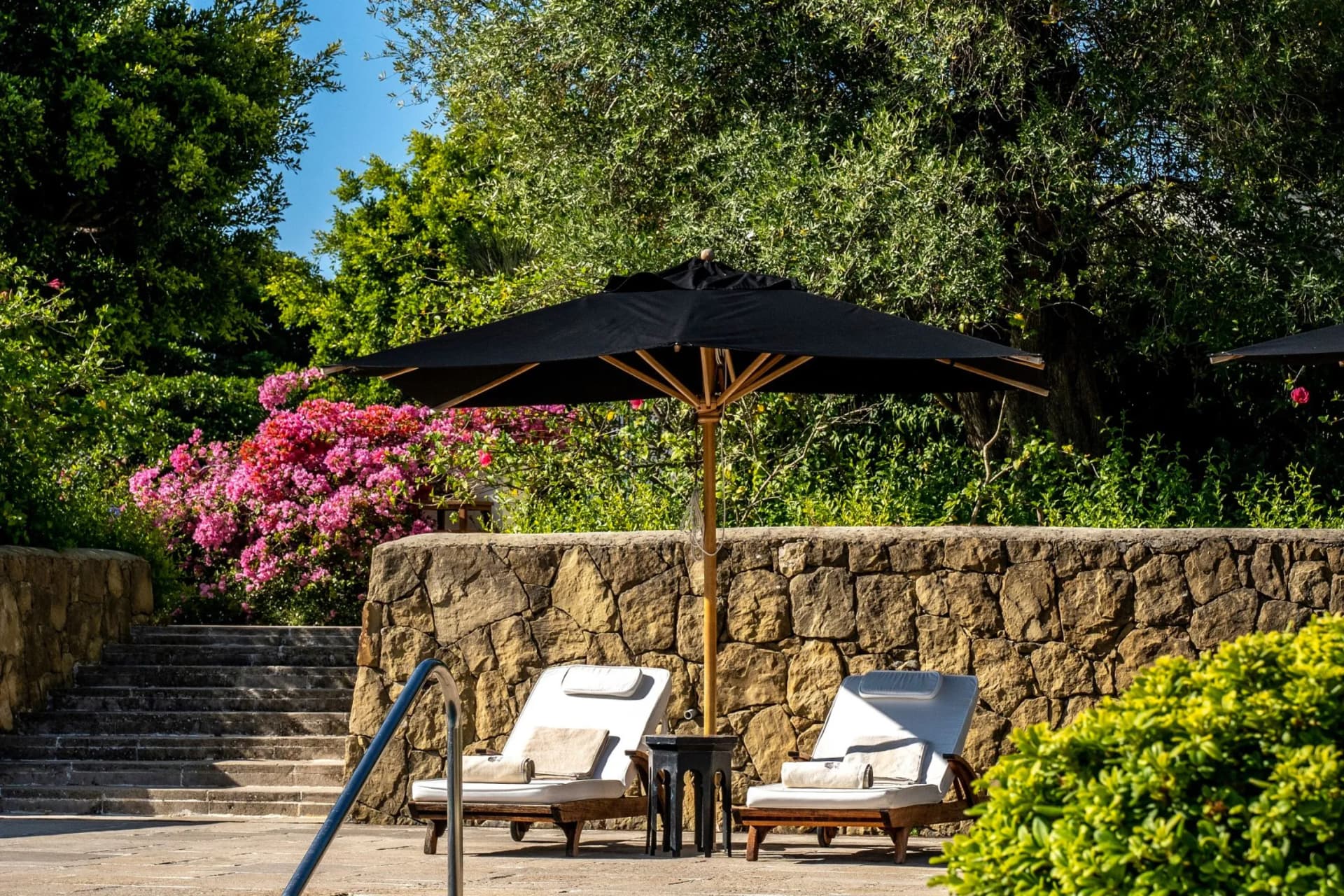 Two white cushioned lounge chairs under a large black umbrella set on a stone patio, with a pink flowering bush and lush green trees behind.