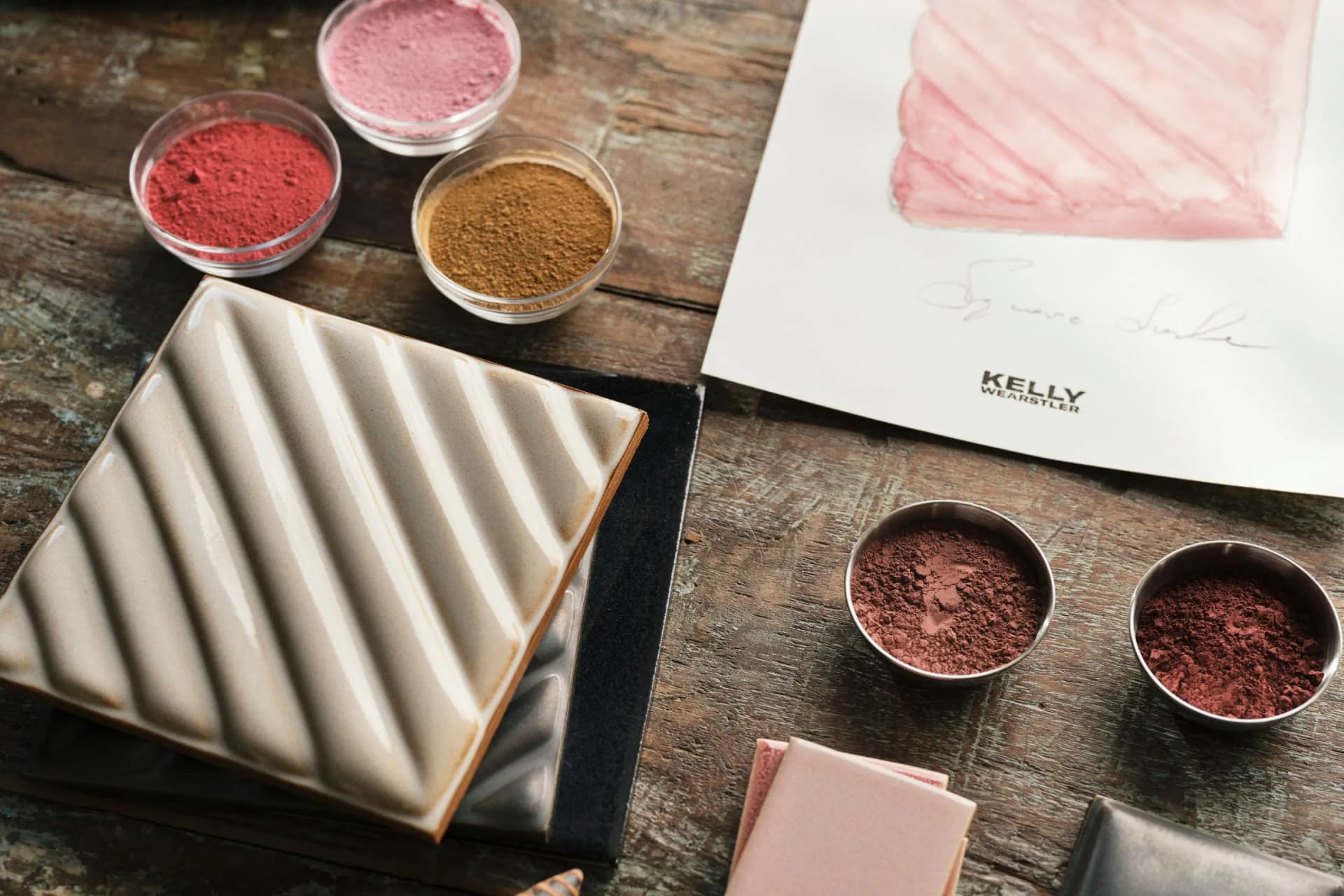 Assorted makeup powders in tins and jars, a large white grooved slab, and a pink swatch sketch on a rustic wooden table.