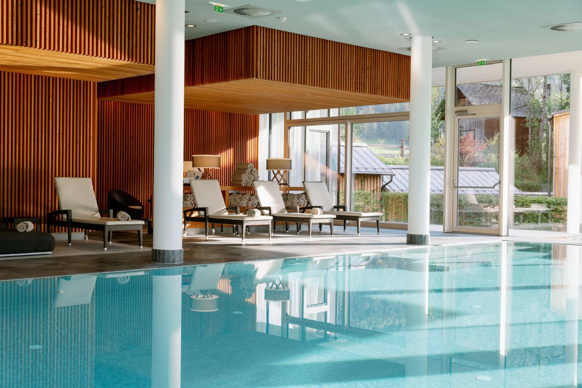Indoor pool with blue water, wood-paneled walls, white columns, and lounge chairs along the pool deck; glass doors open to an outdoor area.