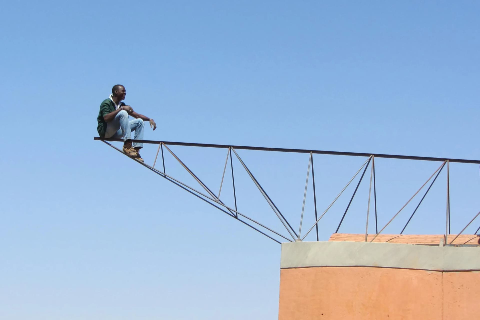 A man sits on a metal beam extending from a rooftop, high above the ground, against a clear blue sky.