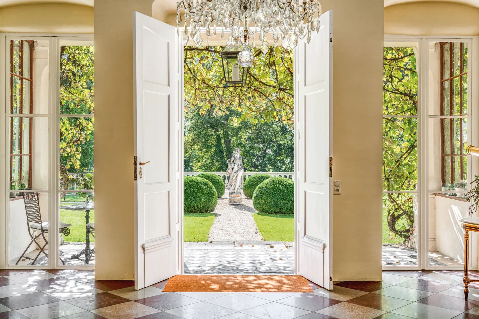 Double white doors open to a sunlit garden with a marble statue, round topiary, and gravel path; crystal chandelier hangs overhead inside.