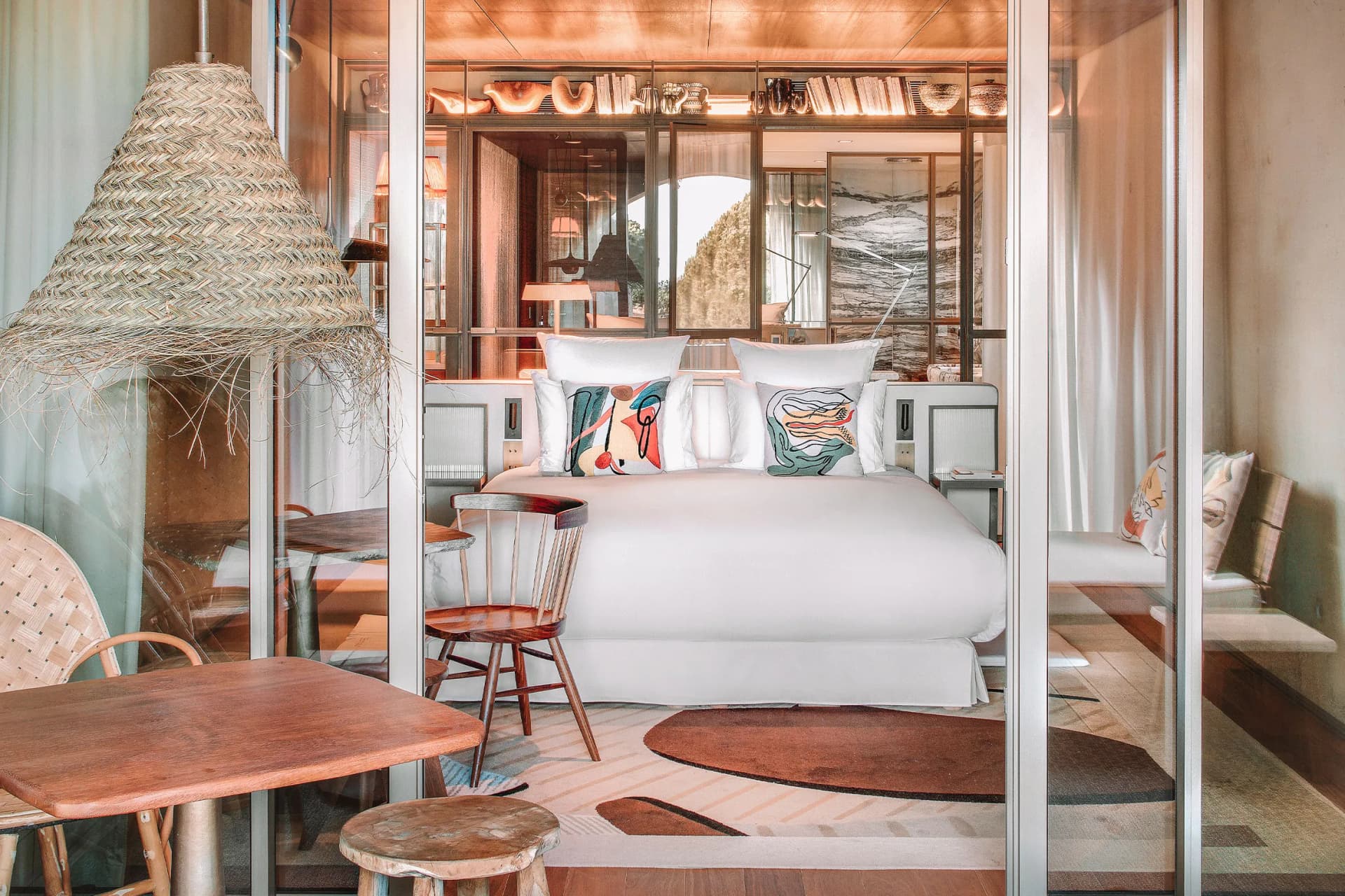 Bright hotel bedroom with white bed, abstract pillows, glass partition, a woven cone lamp on the left, and shelves of decor behind.