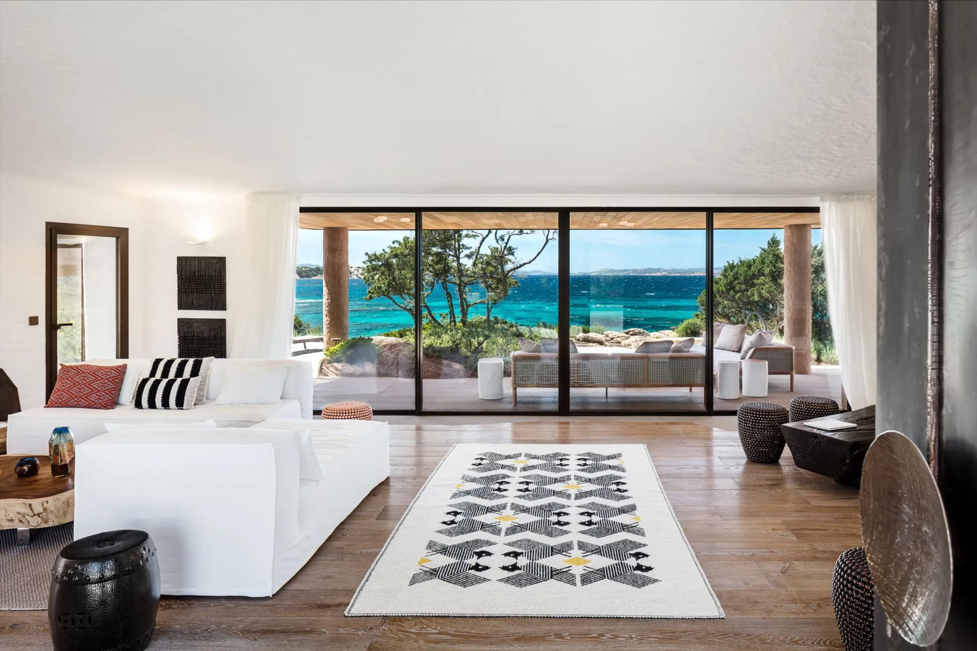 Bright coastal living room with white sofas, a patterned rug, and sliding glass doors to a patio overlooking turquoise sea.