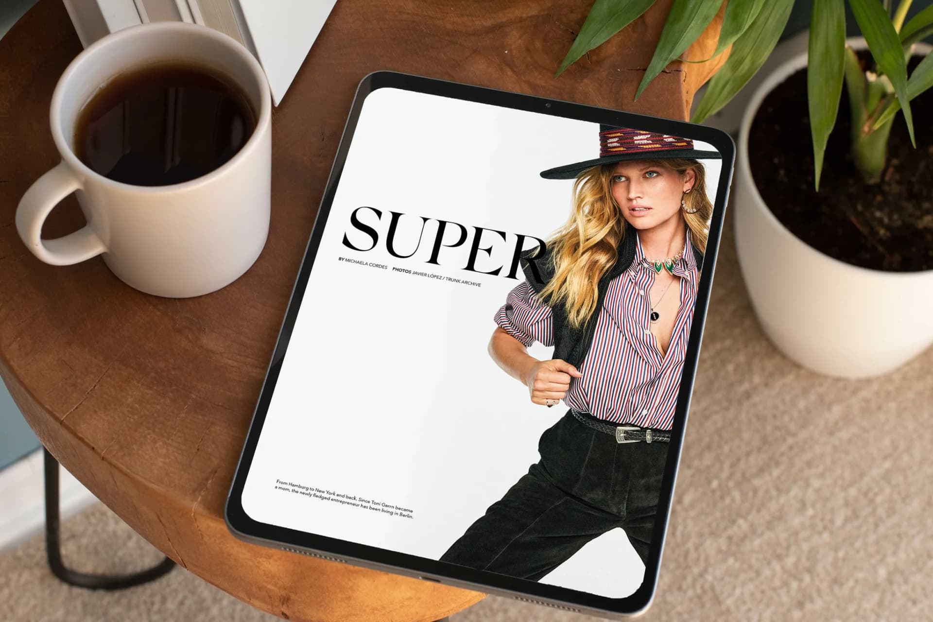 On a tablet resting on a wooden table, the cover of a fashion magazine is visible, featuring a woman with blonde hair and a wide-brimmed hat; next to it are a coffee cup and a plant.