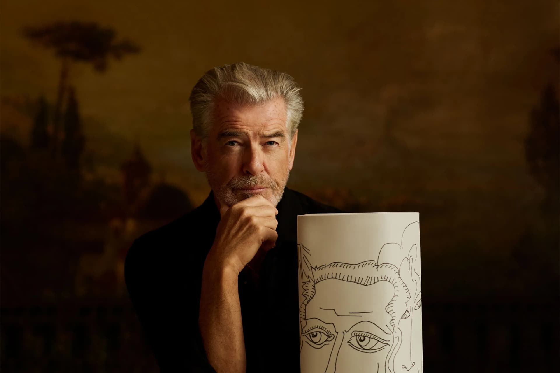 Older man with gray hair and stubble, in dark clothing, holds a tall rolled poster featuring a large drawn face, posing pensively against a dark backdrop.