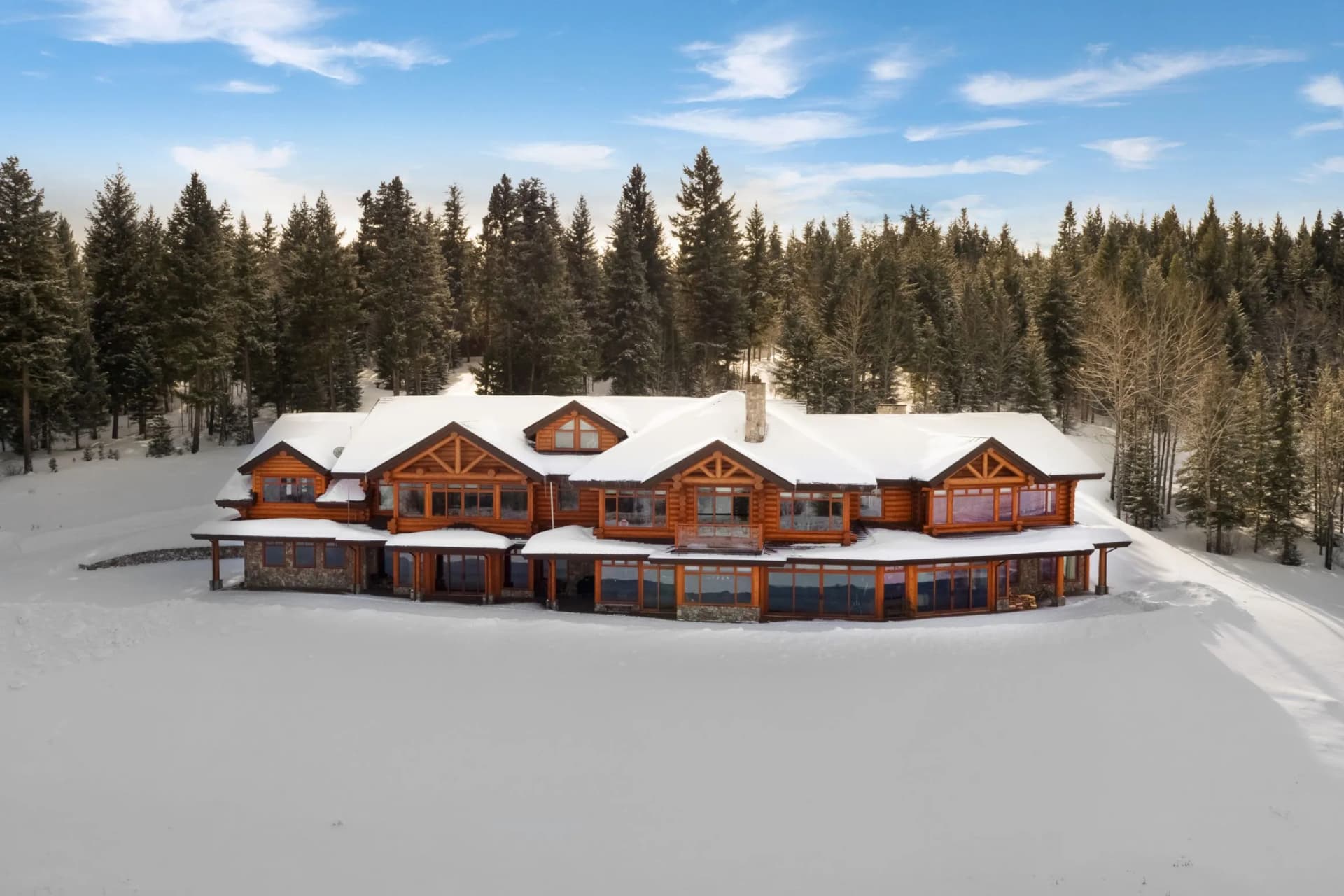 A snow-covered log cabin with large windows stands at the edge of the forest, surrounded by tall evergreen trees beneath a bright blue sky.