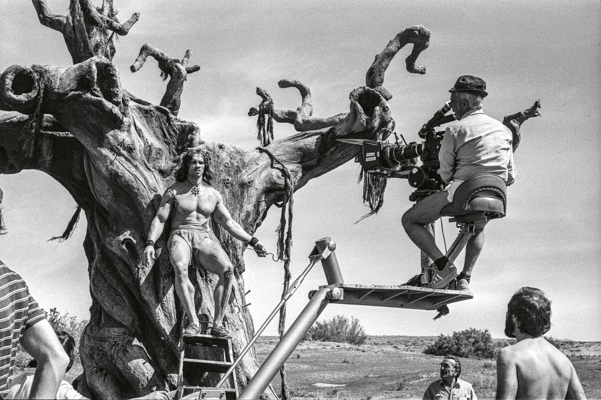 Black-and-white film crew shoots a bikini-clad model perched on a gnarly sculptural tree in a barren landscape; cameraman on a crane watches.