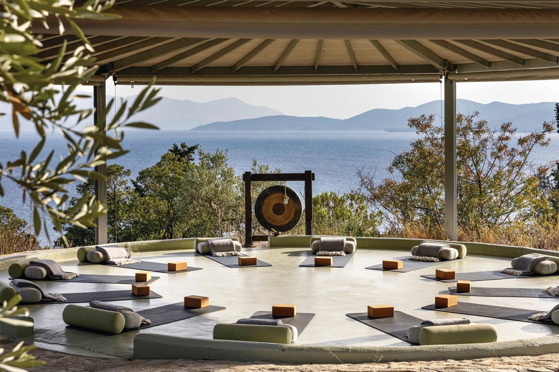 Open-air meditation deck with mats and bolsters arranged in a circle under a pergola, centered by a gong, overlooking the sea and distant mountains.