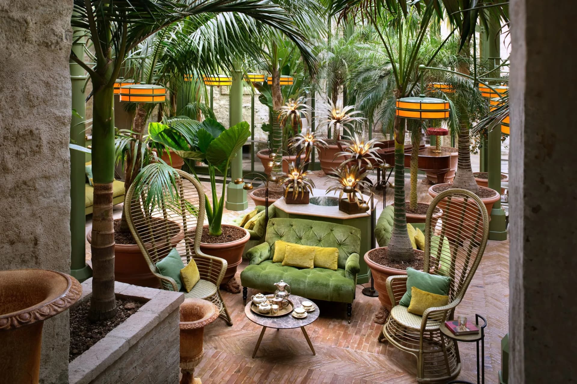 Lush indoor tropical garden seating area with green sofas, wicker chairs, potted palms, and warm orange pendant lamps along green columns.