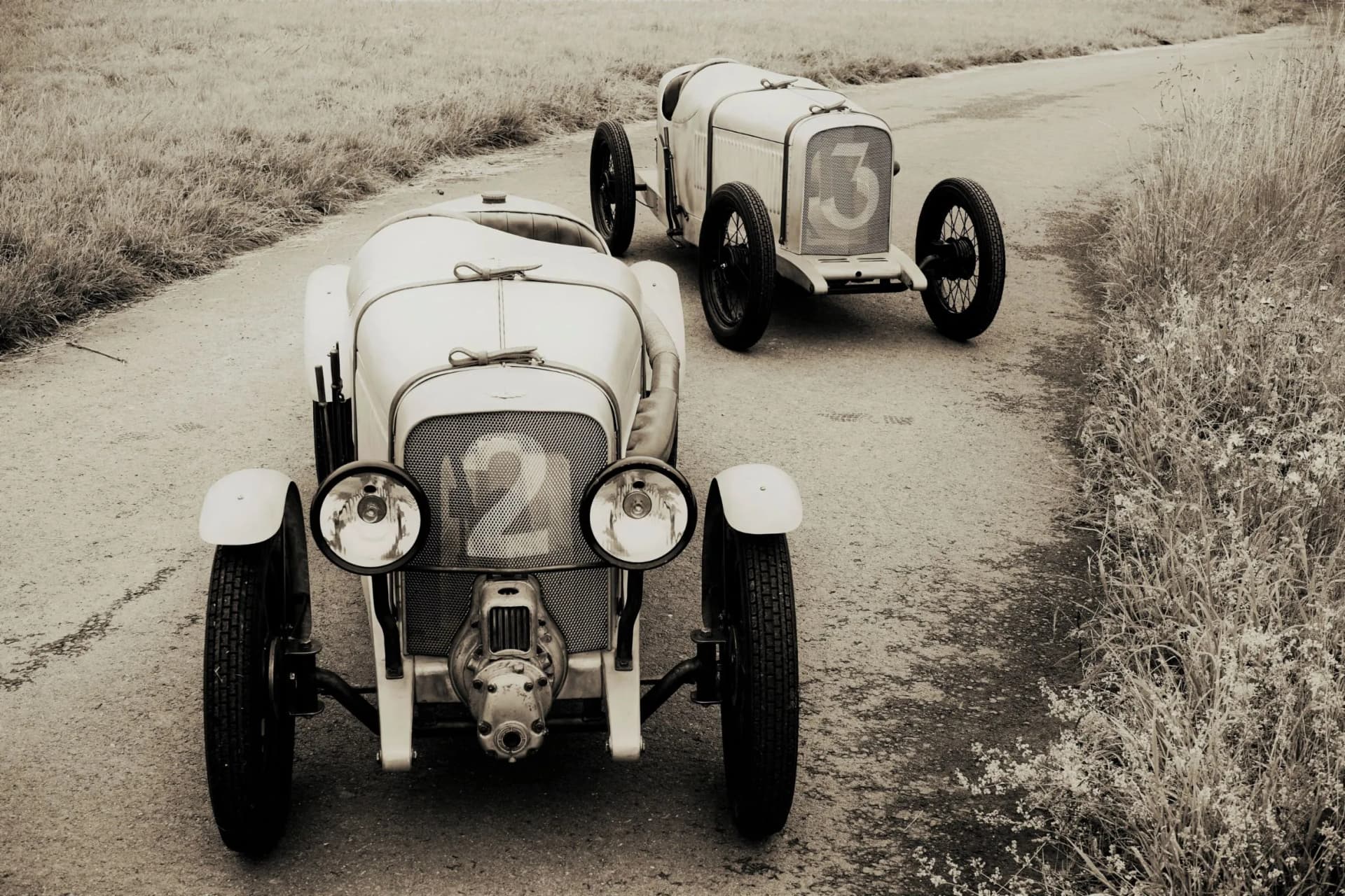 Two vintage open-wheel race cars on a country road, front car marked 2 and rear car marked 3, in sepia tone.