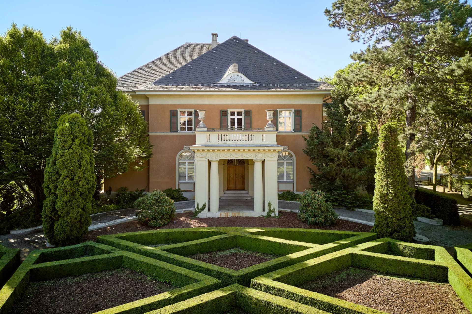 A beige two-storey villa with green shutters and a columned entrance, set amongst formal boxwood hedges and tall trees beneath a blue sky.