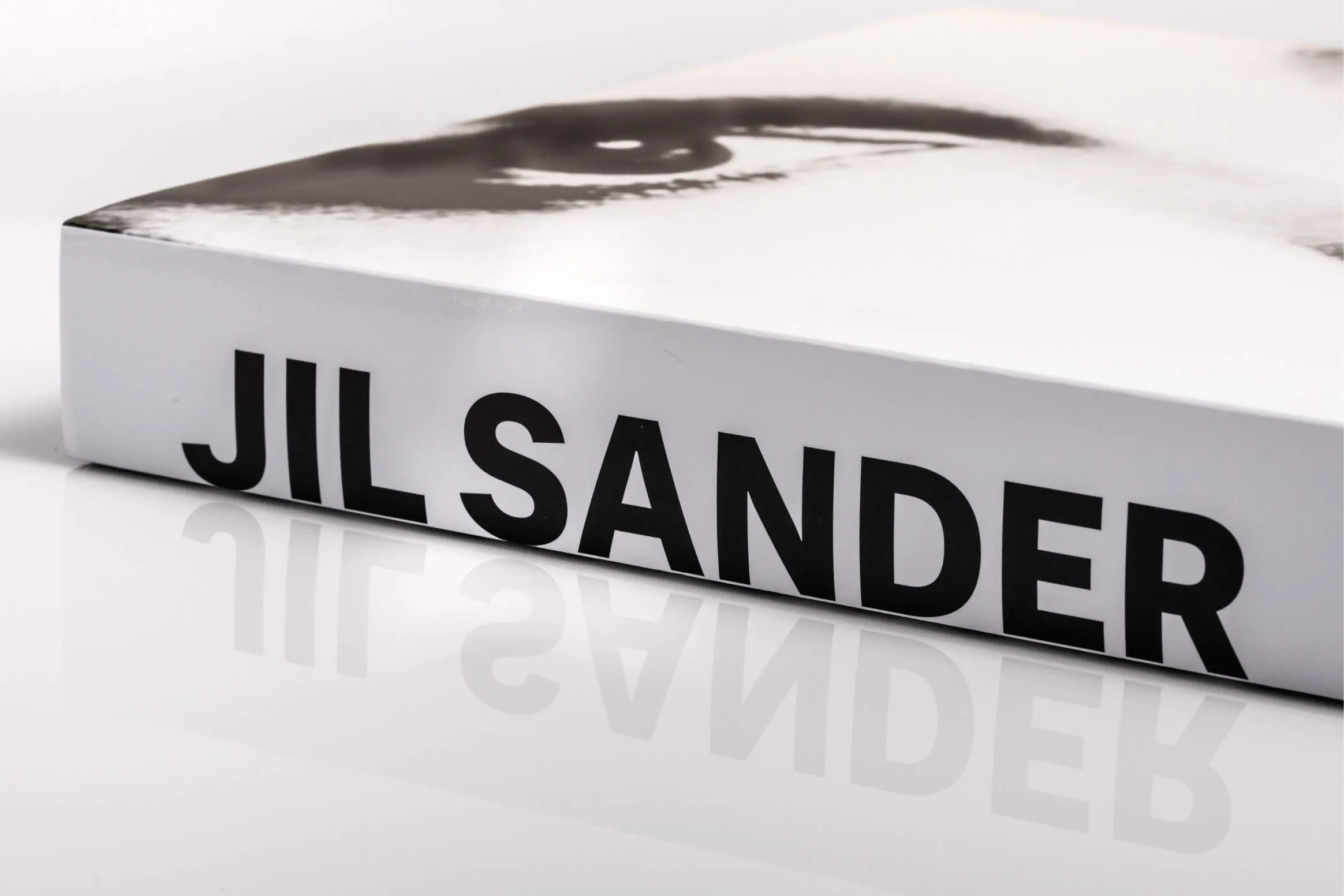 White box with bold JIL SANDER lettering and partial face image on top, reflected on a glossy surface.