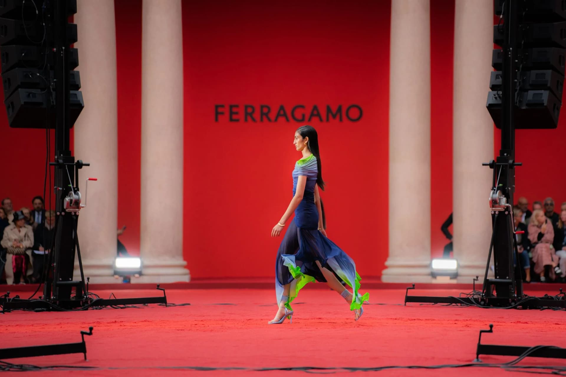 Model walks a red runway in a blue flowing dress with lime-green heels, flanked by columns and an audience, Ferragamo backdrop.