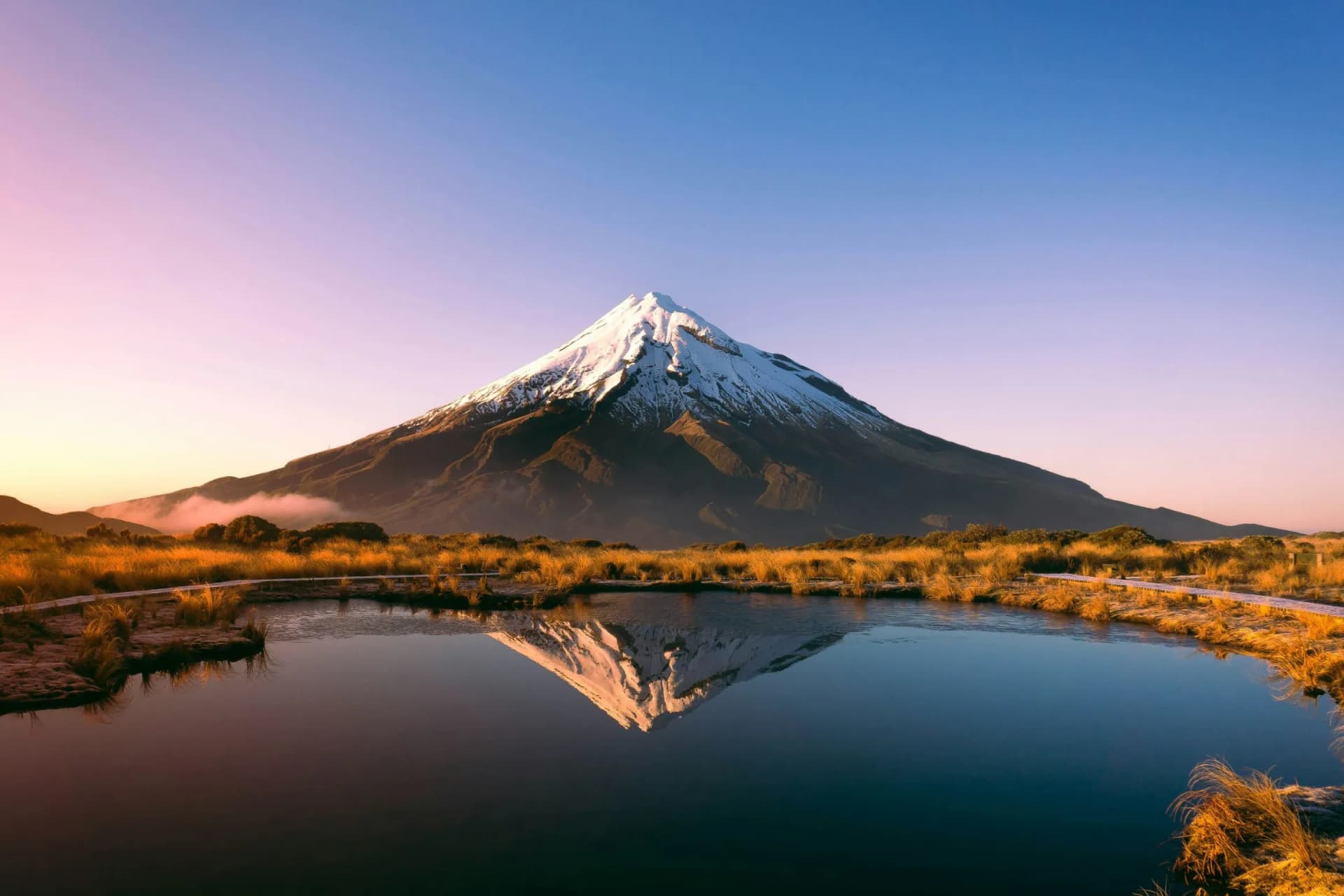 Snow-capped mountain mirrored in a calm lake, with golden grasses along the shore and a pastel sunrise lighting the sky.