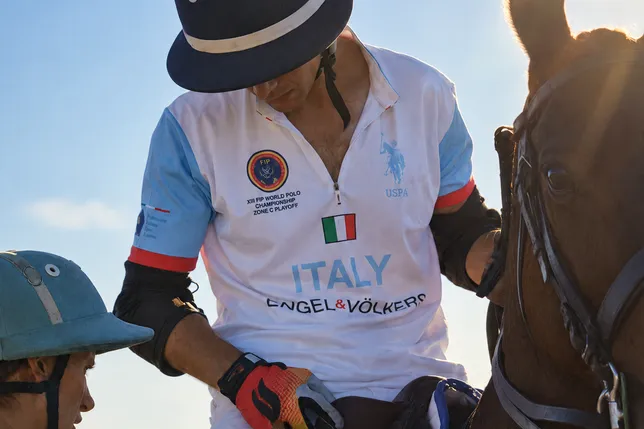 Polo player in an Italy uniform sits on a horse wearing a dark helmet as a teammate tightens straps; another blue helmet rests nearby against a sunny sky.