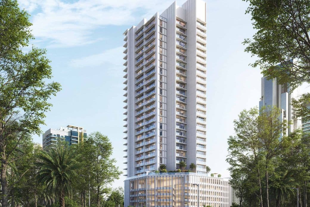 Tall modern apartment building surrounded by trees, with a clear blue sky and scattered smaller buildings in the background.